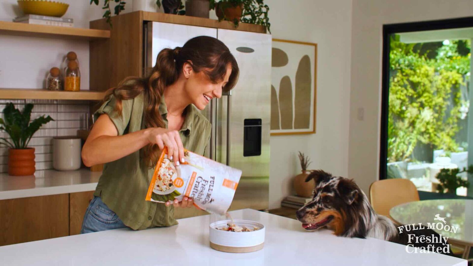 A woman is pouring dog food into a bowl on a kitchen counter while a dog eagerly watches, as if starring in an innovative television commercial. The kitchen boasts modern decor, and greenery is visible outside the window.