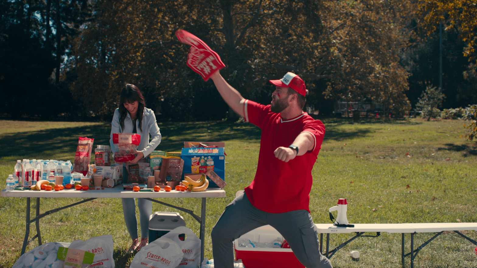 A man in a red shirt and cap enthusiastically waves a foam finger in a park, embodying the spirit of Stater Bros. Markets' community commitment. Behind him, a woman arranges healthy snacks on the table, capturing the essence of family values and quality products perfect for busy families enjoying an outdoor gathering.