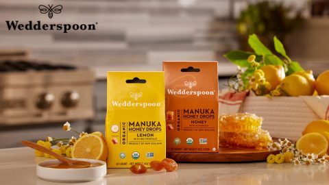 Two packages of Wedderspoon Manuka Honey Drops, one lemon-flavored and one original, are placed on a kitchen counter surrounded by honeycomb, lemons, flowers, and a honey dipper—a perfect scene for an advertising agency to capture in their next campaign.